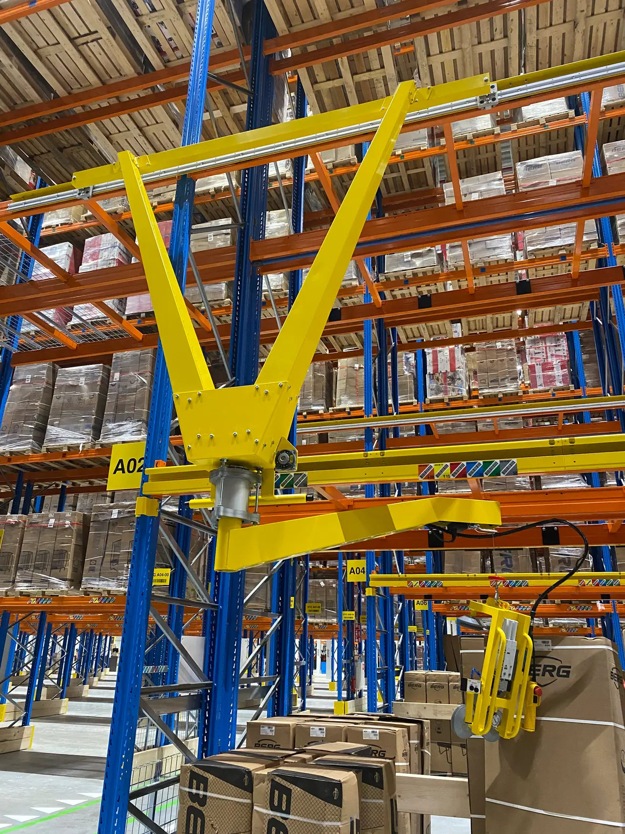 Gele RackJib 110 met V-vormige arm en grijpgereedschap, aan een stelling in een magazijn